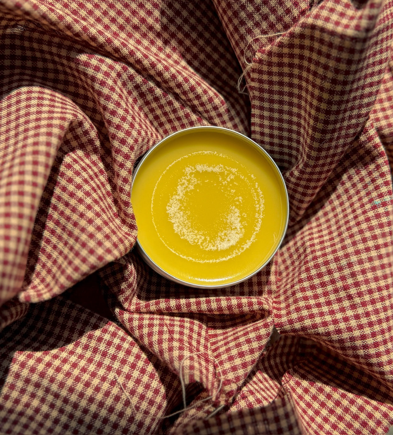 An open tin of golden Soothe Salve resting on red gingham fabric