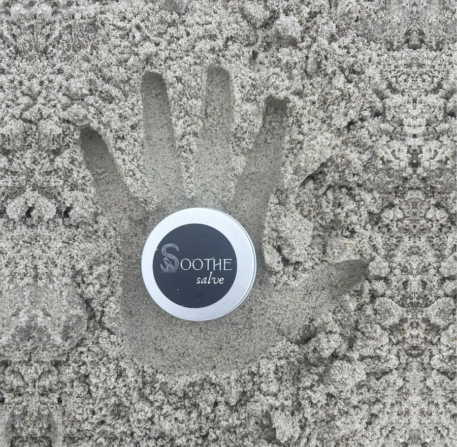A round tin of Soothe Salve placed in the center of a handprint made in sand, with the brand name 'SOOTHE salve' visible on the lid.